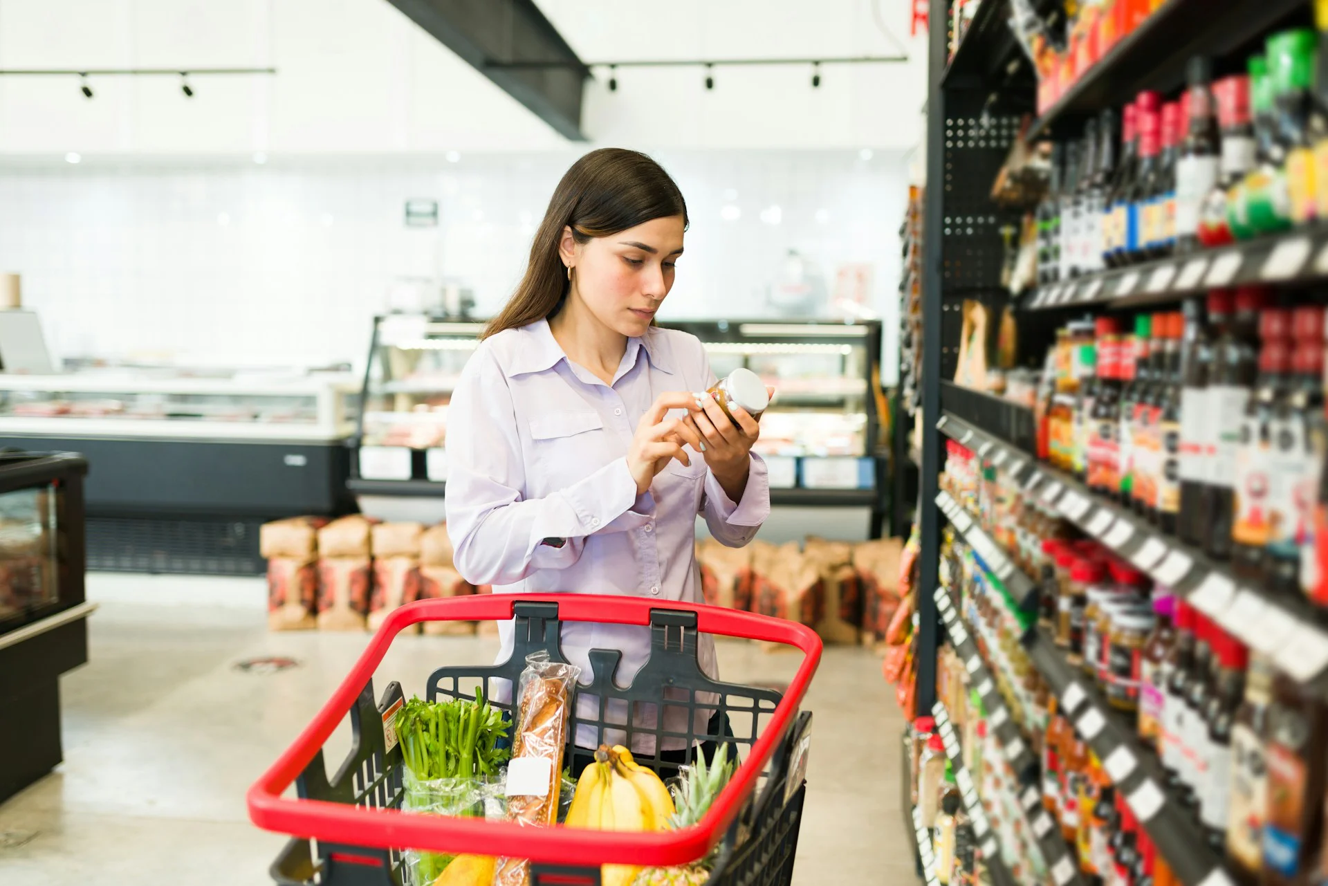 Una donna legge l’etichetta nutrizionale di un prodotto confezionato al supermercato, con il carrello pieno di verdura fresca e prodotti da scaffale